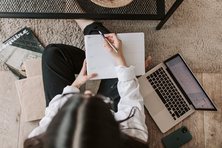 Top view of anonymous woman in casual wear sitting on floor with laptop and smartphone and creating plan on notebook while googling best mortgage broker Perth in modern living room