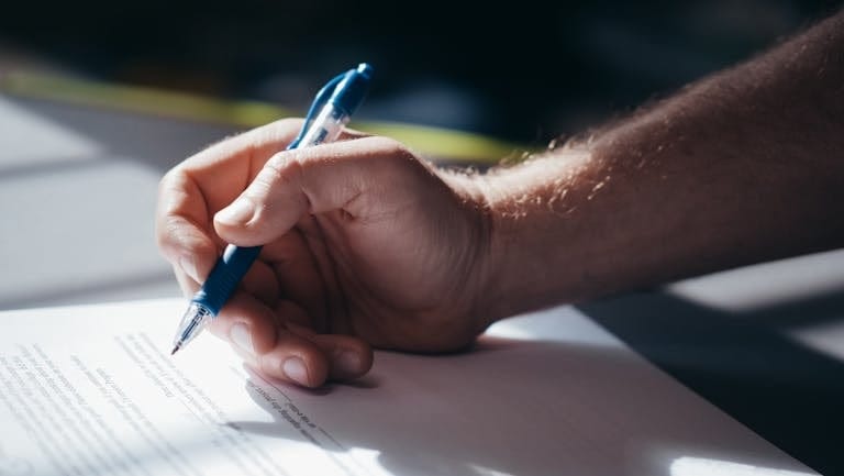 Detailed shot of a hand holding a blue pen while signing a document in accordance with best interest duty for mortgage brokers.