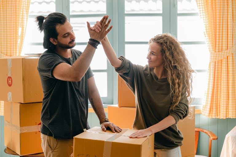 Happy woman in casual wear standing near heap of cardboard boxes and giving high five to ethnic boyfriend with ponytail showing agreement while looking at each other after they used the FHSS scheme and super to help buy a home