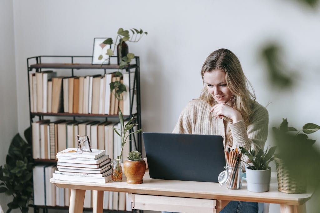 Interested young self employed woman on notebook researching home loans for self employed at table with potted plants against bookshelves in house on light background