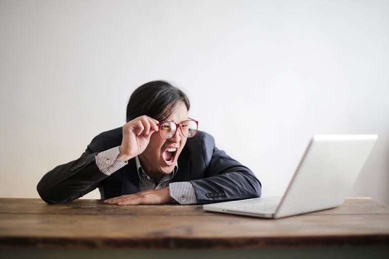 Modern Asian man in jacket and glasses looking at laptop and screaming with mouth wide opened on white background when he finds out about settlement delays from his mortgage broker perth
