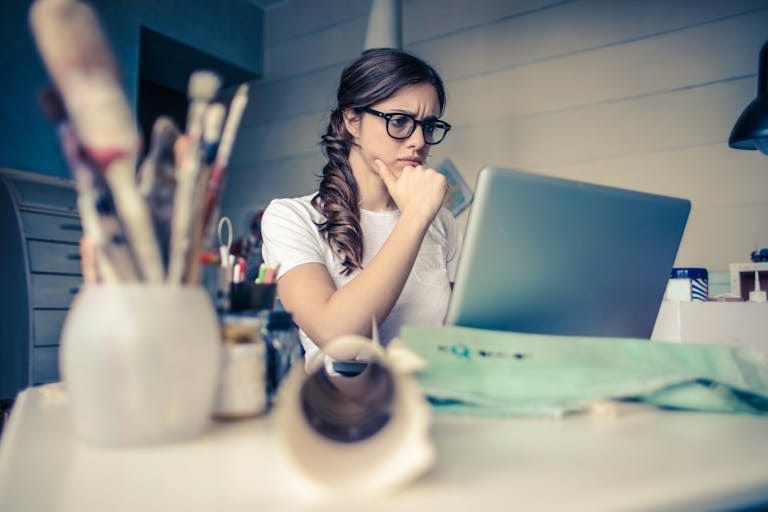Young woman with glasses deeply focused on a laptop surrounded by art supplies in a home office as she looks for a mortgage broker perth to help with near prime lending