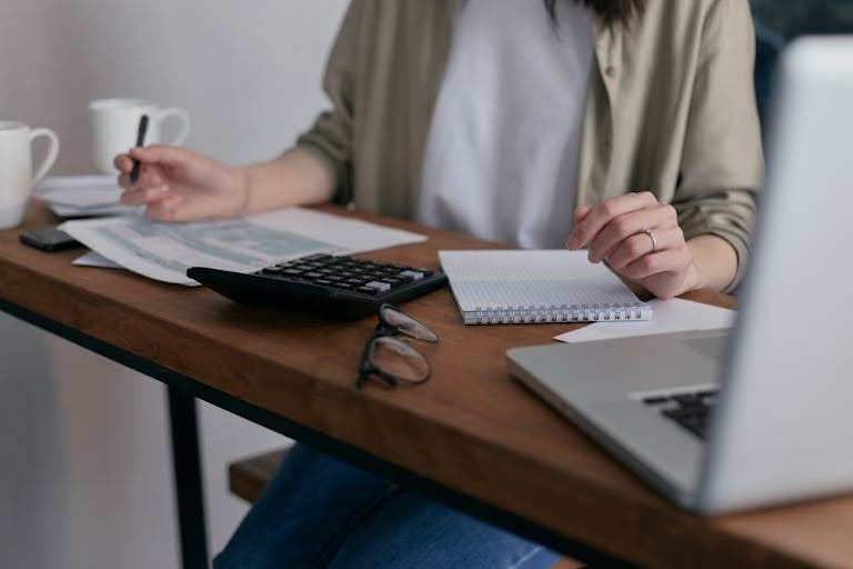 A woman calculates her home loan deposit, using a laptop and calculator on a wooden desk after consulting her perth mortgage broker