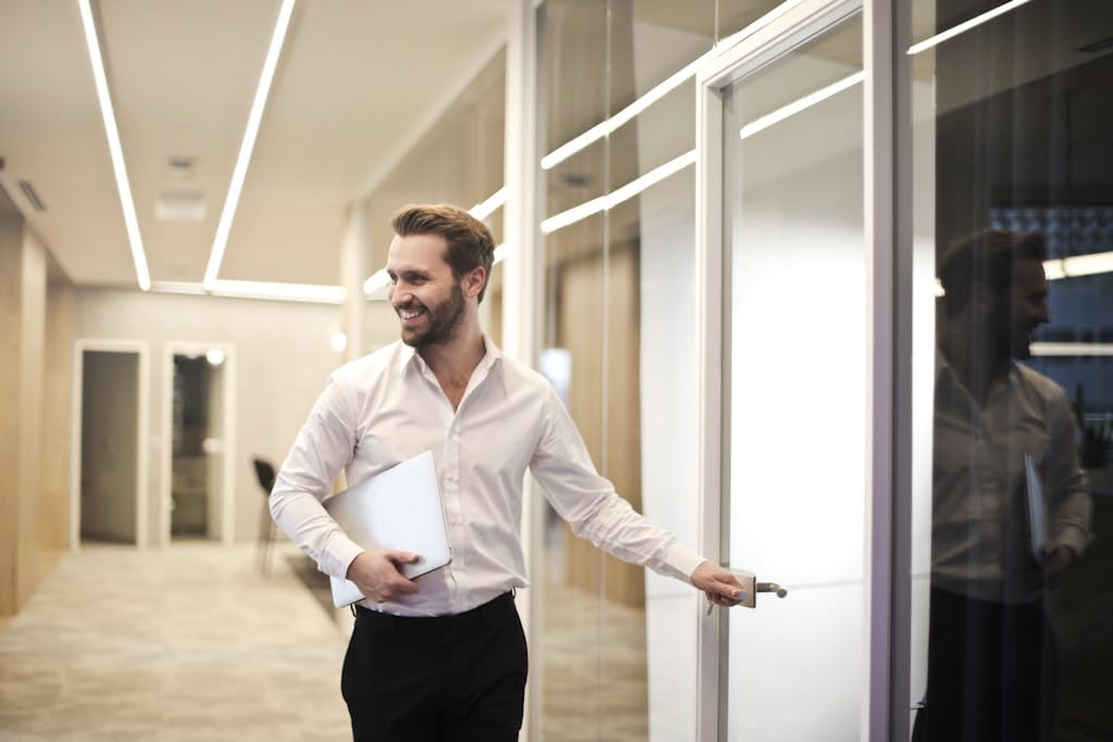 A smiling man holds a laptop while standing in a modern office hallway, heading to meeting with perth mortgage broker to discuss commercial finance.