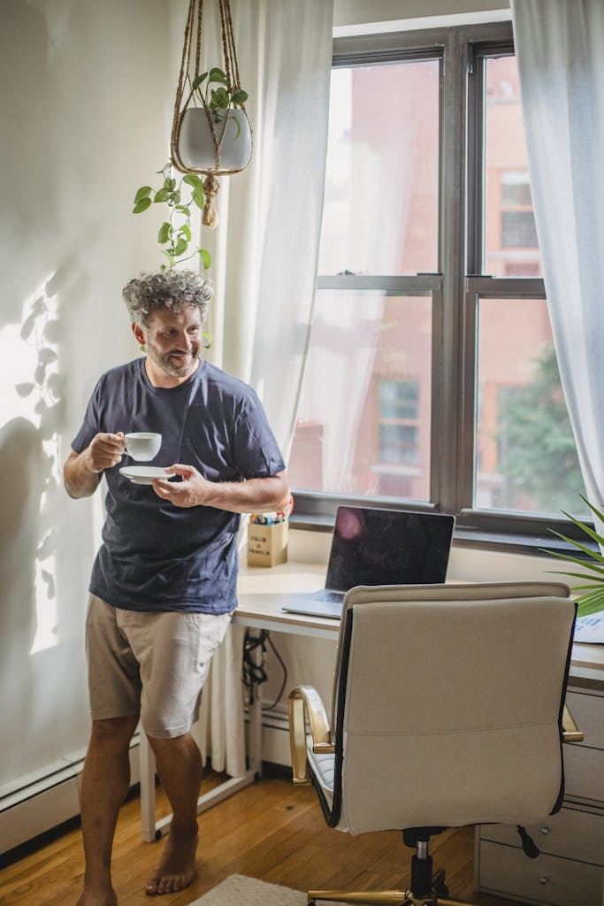 Happy adult bearded man in home outfit leaning on table with modern laptop and drinking coffee during break in freelance work on sunny day after speaking with his perth mortgage broker about alternative lending for self employed