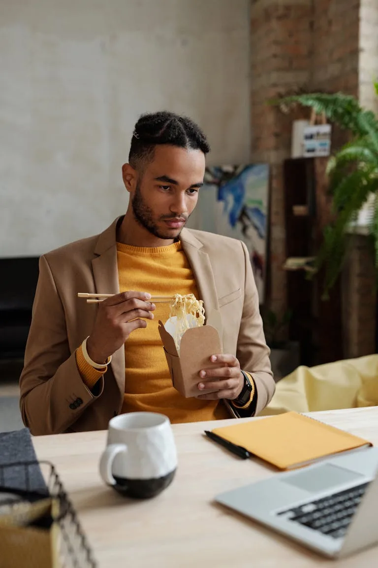 Young man in casual blazer eating noodles with chopsticks while working at a desk researching first homebuyer journey in Perth.