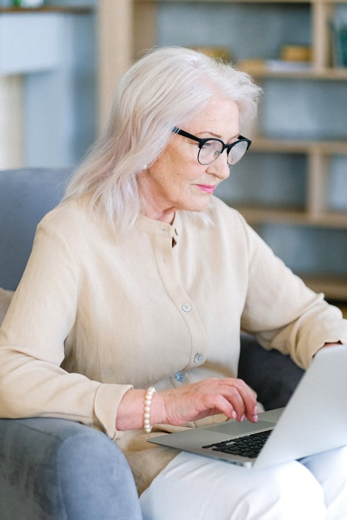 Elderly woman with glasses typing on a laptop in a cozy home setting, planning for retirement with property investment perth