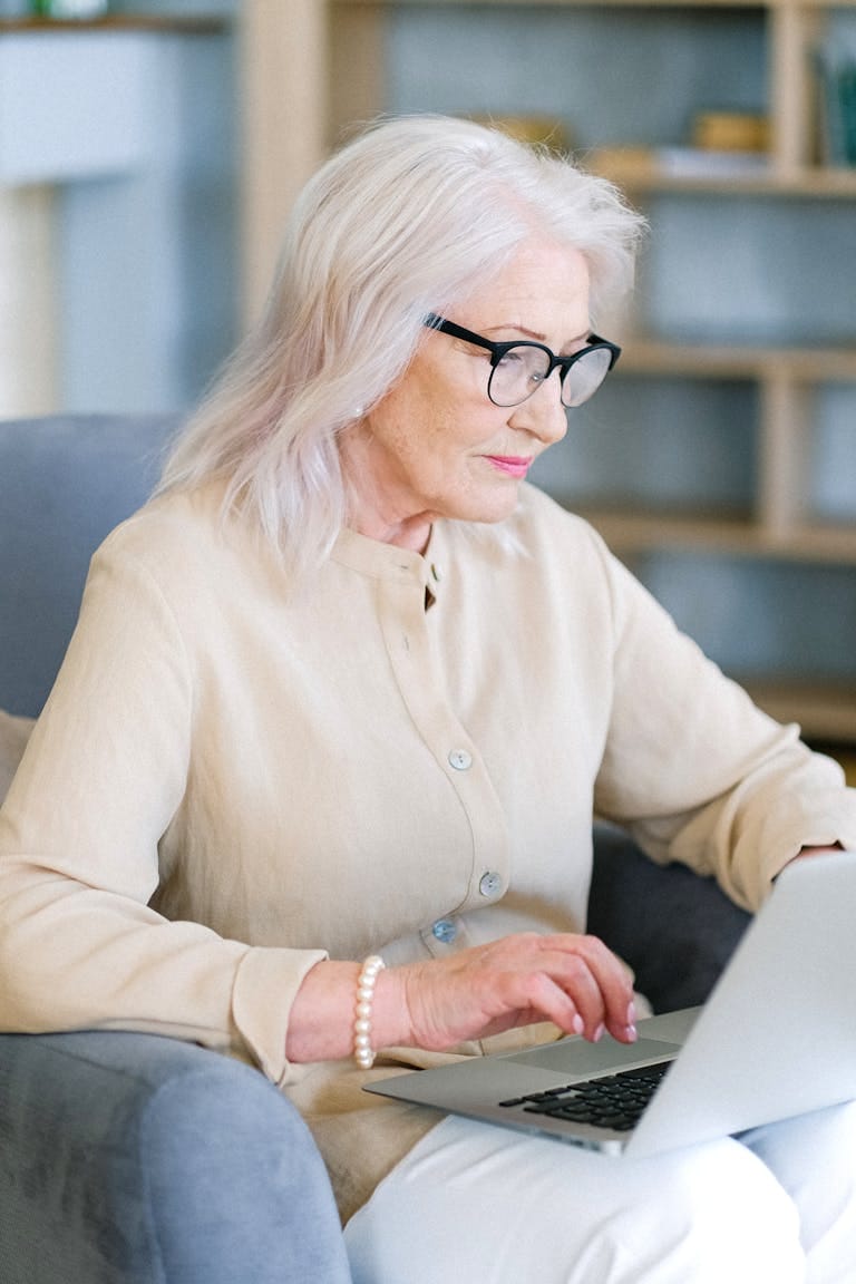 Elderly woman with glasses typing on a laptop in a cozy home setting, planning for retirement with property investment perth