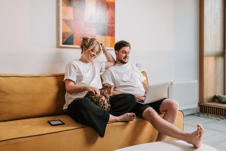 A couple sitting relaxed on the sofa with their cat, engaging in remote work using a laptop as they work to become mortgage free in perth
