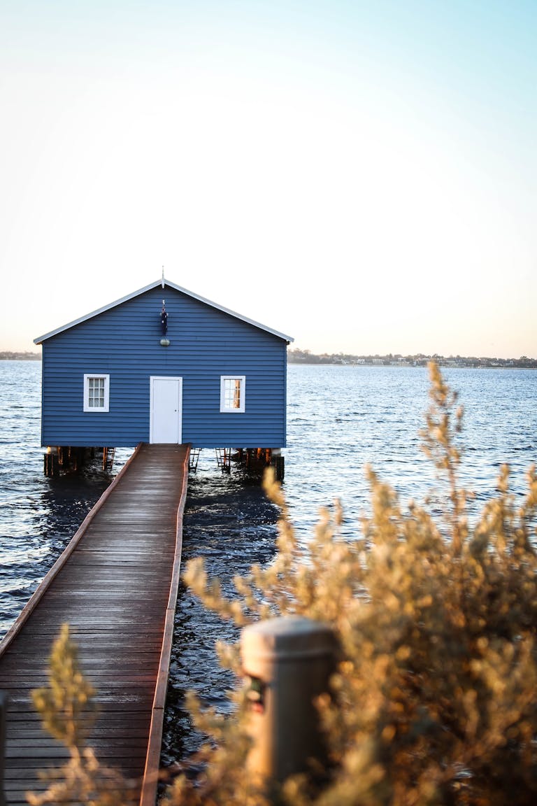 A tranquil scene of a blue boathouse on Swan River, with a wooden walkway and soft lighting as people question perth property market in 2026