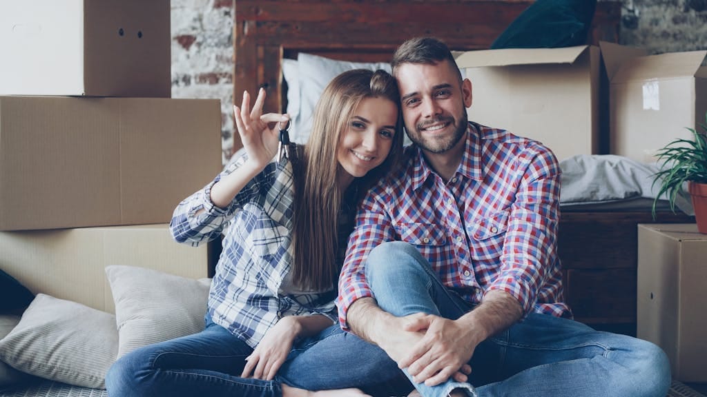 Smiling couple in first home holding keys, surrounded by moving boxes after thinking it was impossible to buy a first home
