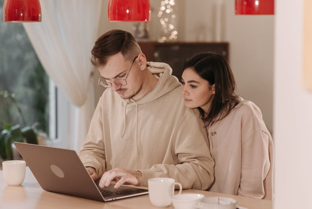 Young couple in hoodies using a laptop at home with coffee cups, as they research refinancing options